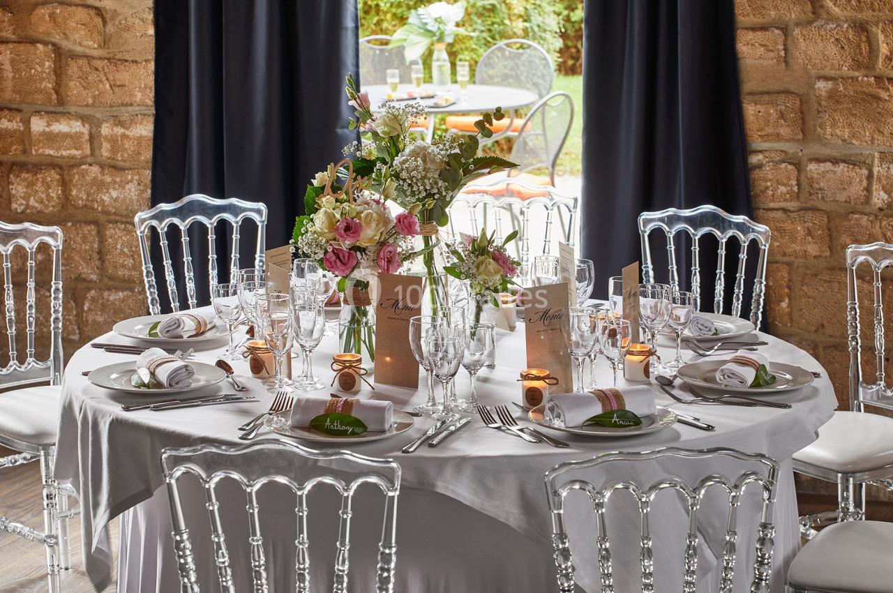 Table élégamment dressée avec nappes blanches, fleurs au centre, menus et chaises transparentes dans une salle en pierre.