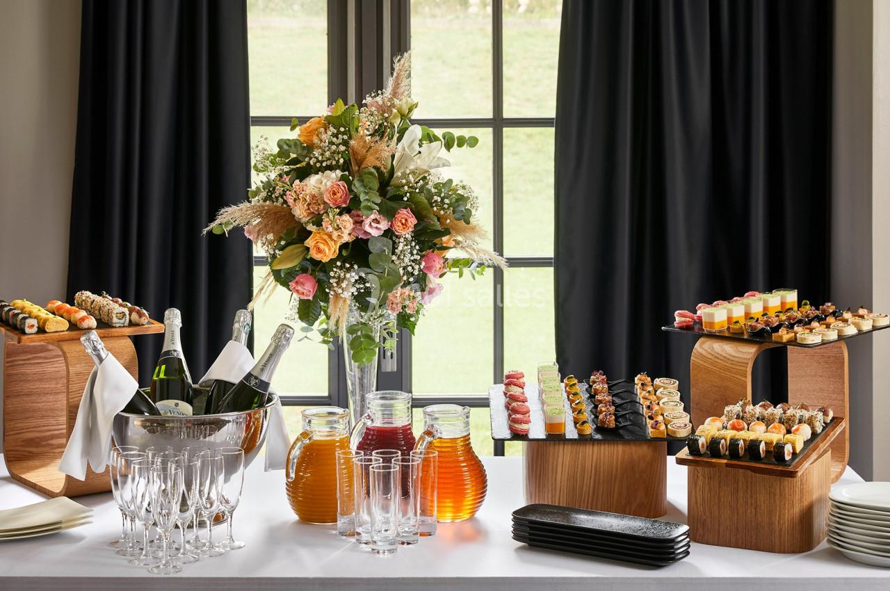 Table dressée avec des amuse-bouches variés, un bouquet de fleurs, des boissons et des verres devant une fenêtre.