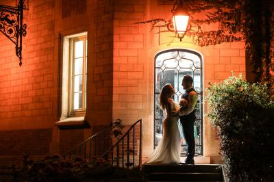 Un couple en tenue de mariage se tient devant une porte illuminée, entouré de briques et de végétation.