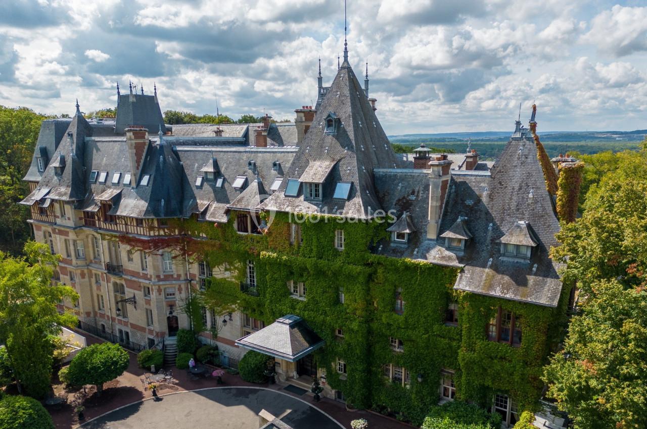 Vue aérienne d'un château couvert de lierre, entouré de végétation, sous un ciel partiellement nuageux.