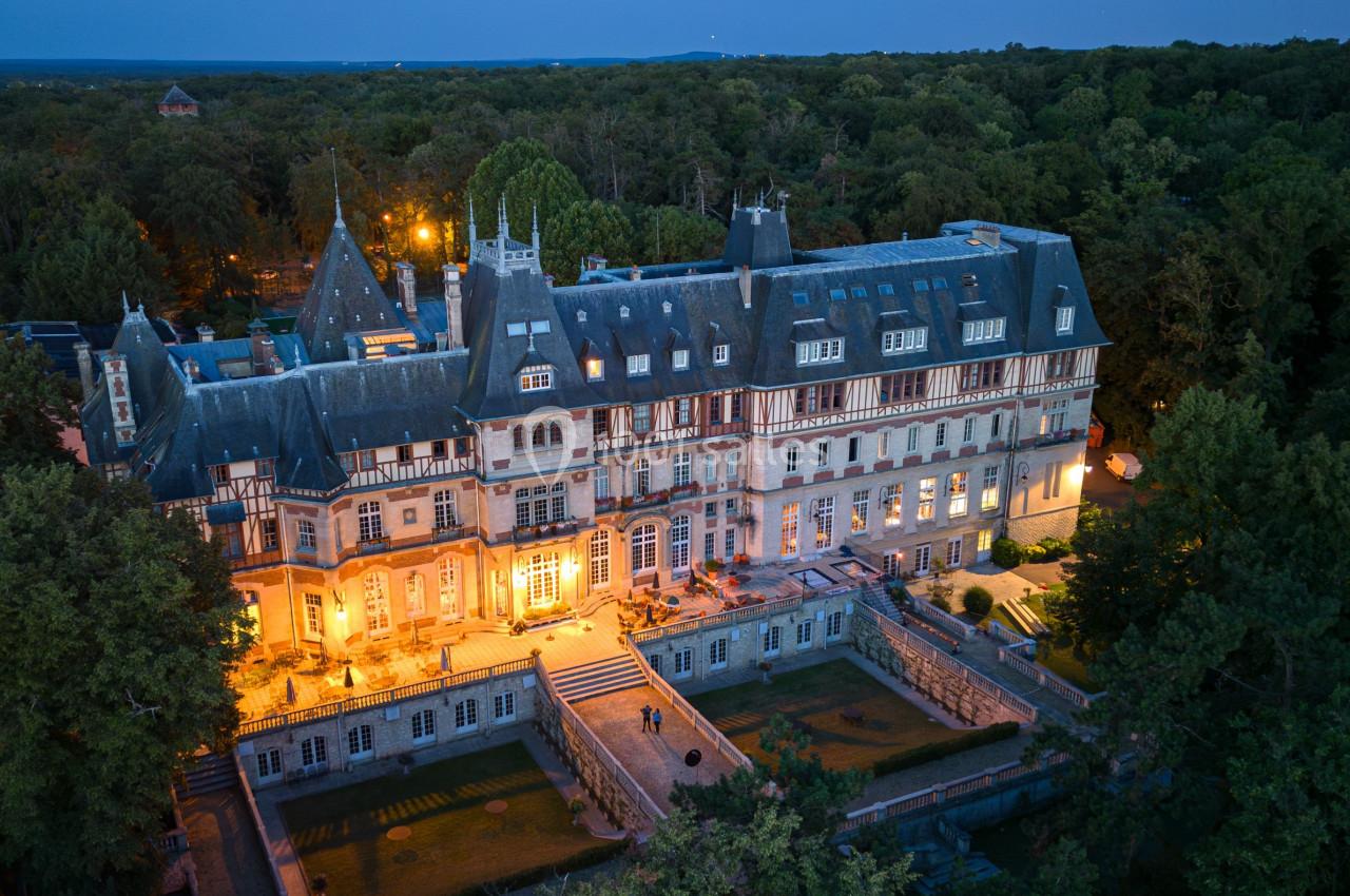 Vue aérienne d'un grand château éclairé au crépuscule, entouré de forêt dense.