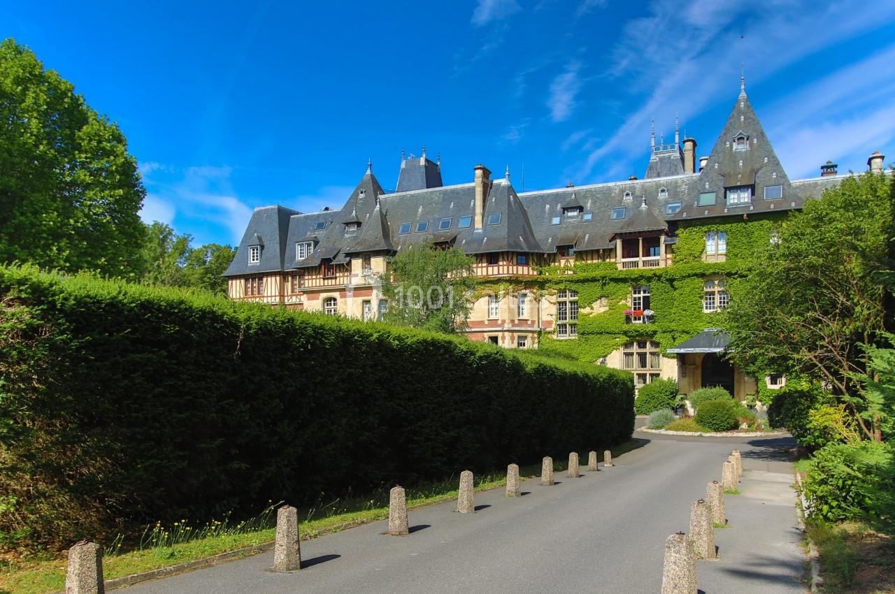 Allée bordée de haies menant à un grand bâtiment couvert de lierre avec des toits en ardoise sous un ciel bleu.