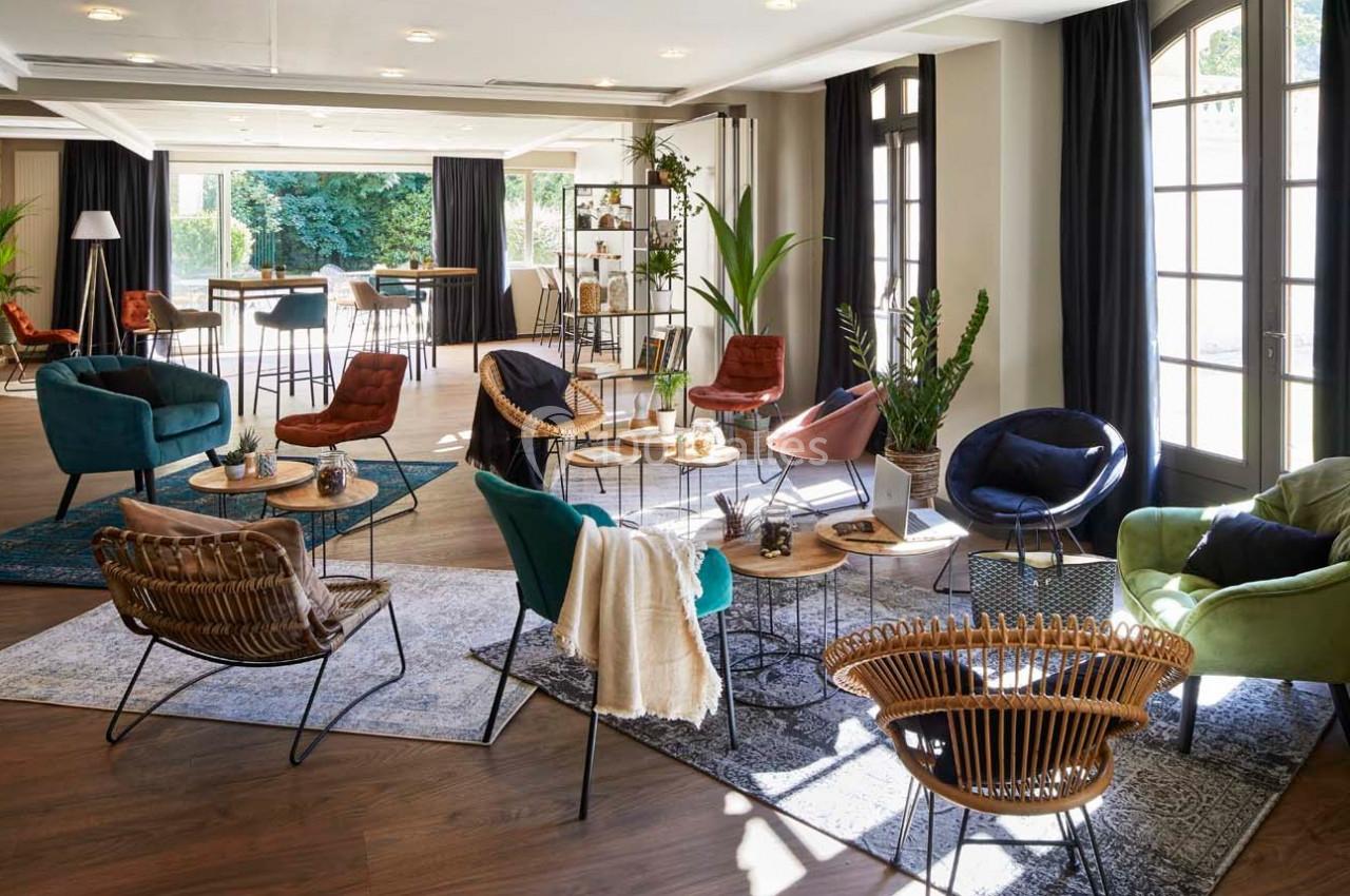 Salle lumineuse aménagée avec des fauteuils variés, des tables basses et des plantes, donnant sur un espace extérieur…