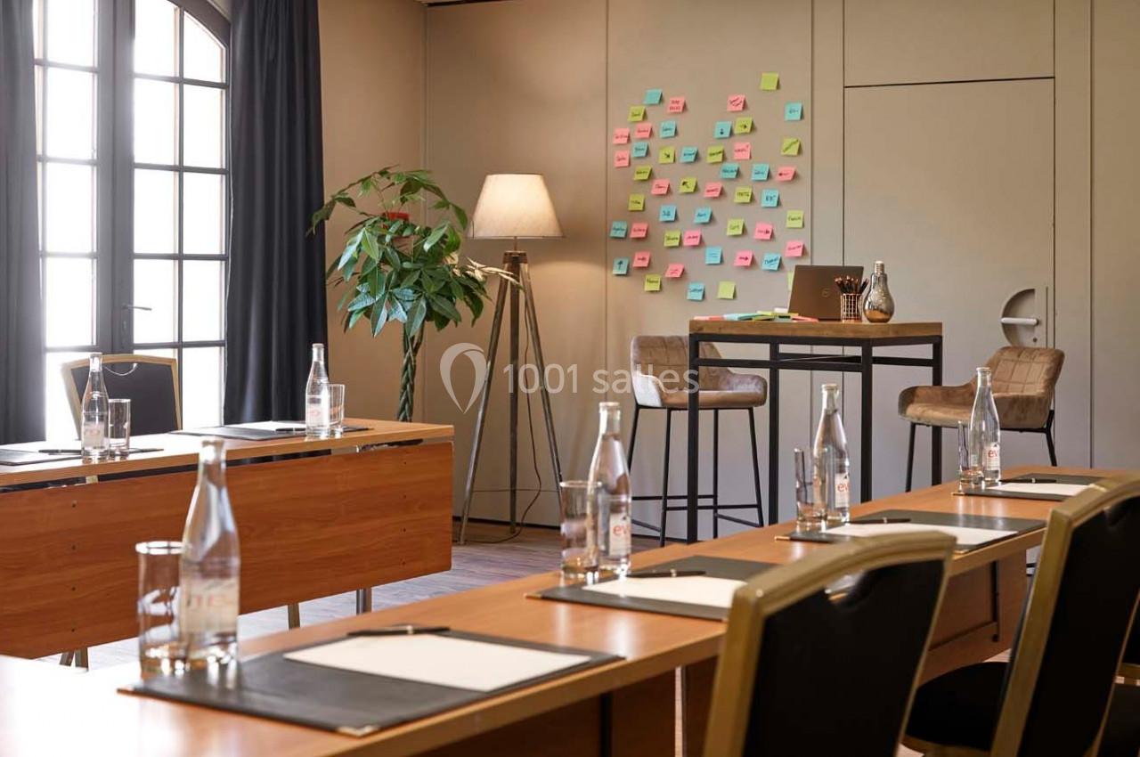 Salle de réunion lumineuse avec tables en bois, chaises noires, tableau couvert de post-it colorés et bouteilles d'eau.