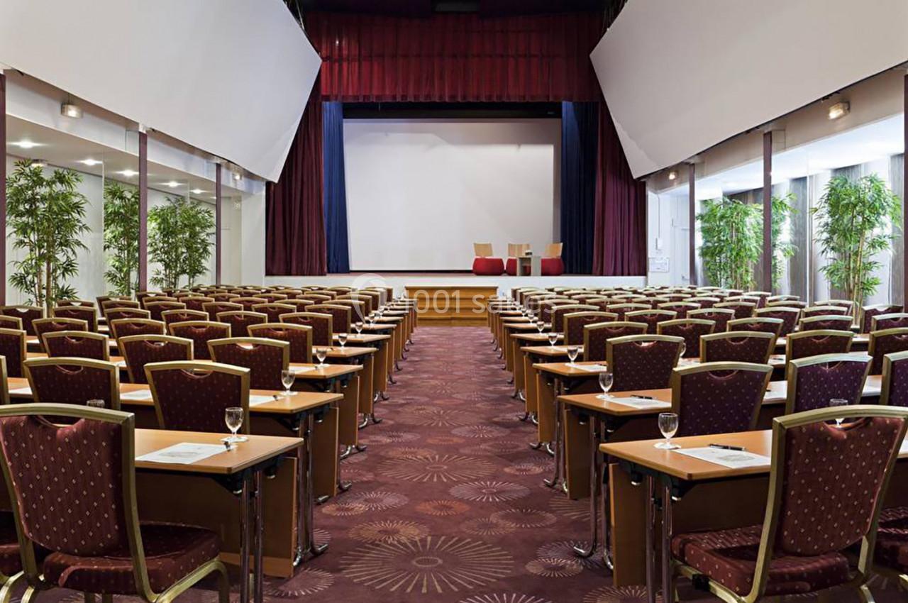 Salle de conférence avec rangées de chaises et tables alignées face à une scène équipée d'un écran blanc.