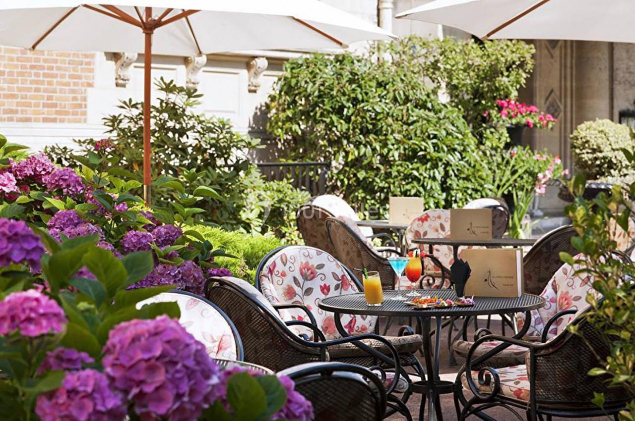 Terrasse fleurie avec tables et chaises en fer forgé, boissons servies sur une table, parasols et verdure environnante.