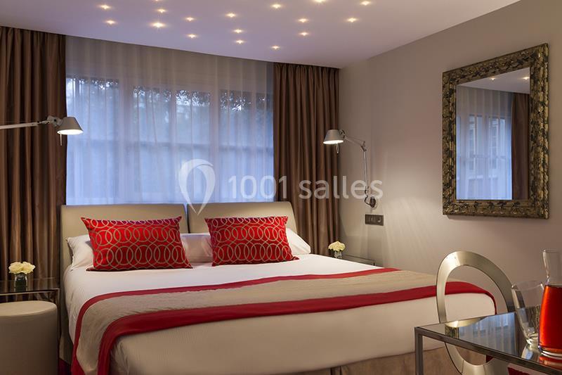 Chambre moderne avec lit double, coussins rouges, éclairage tamisé, grande fenêtre et miroir encadré sur le mur.