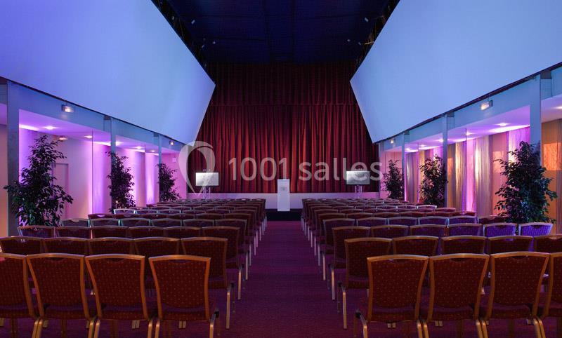 Salle de conférence avec des rangées de chaises vides, un podium central et des écrans, éclairée par des lumières violettes.