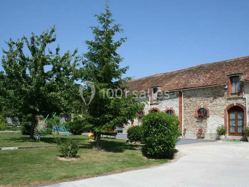 Ferme en pierre avec toit en tuiles, entourée d'arbres, pelouse et une balançoire dans un jardin ensoleillé.