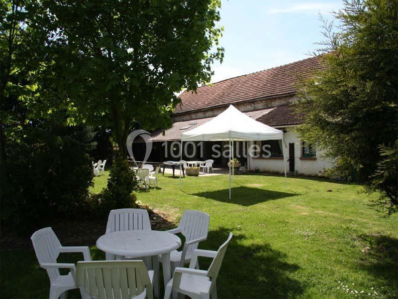 Jardin avec tables et chaises blanches en plastique, une tente blanche et une maison en arrière-plan sous un ciel dégagé.