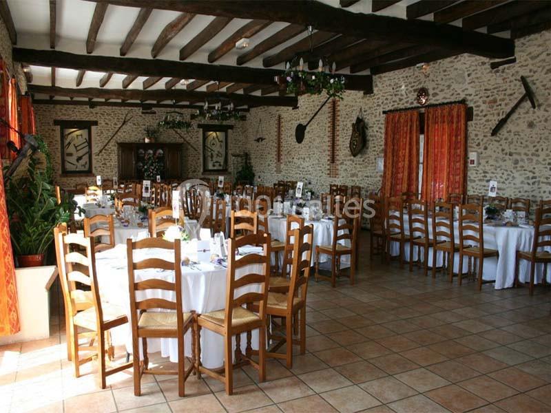 Salle de réception rustique avec murs en pierre, poutres apparentes, tables rondes dressées et chaises en bois.