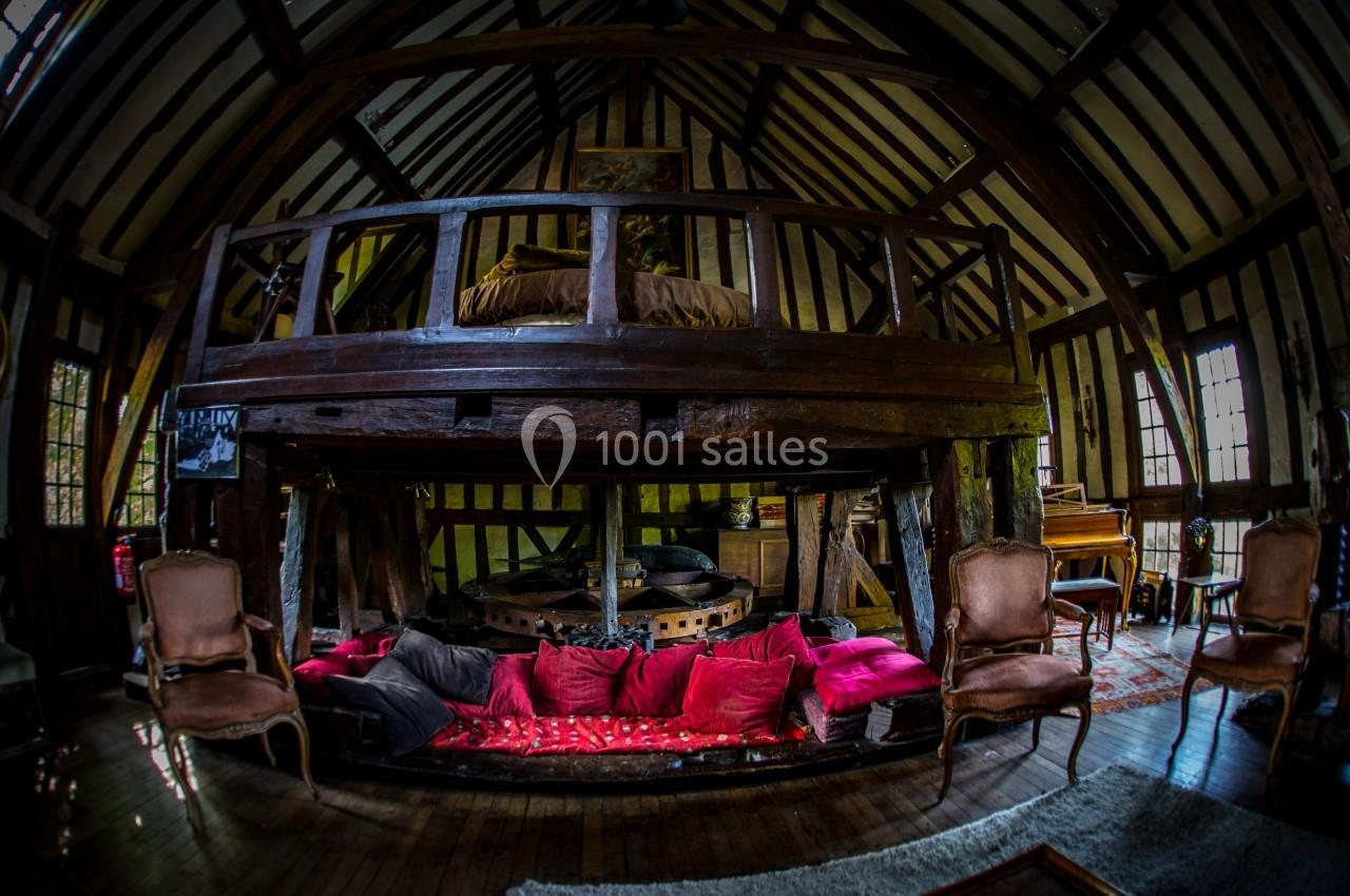 Intérieur d'un moulin rénové avec poutres apparentes, mezzanine en bois, fauteuils anciens et coussins colorés.