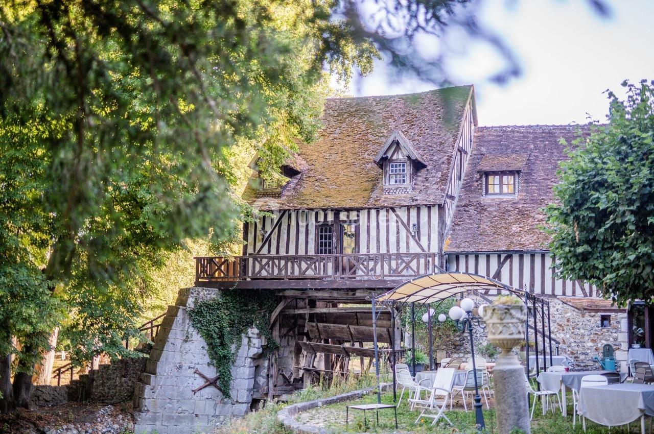 Maison à colombages avec toit en chaume, entourée de verdure, avec terrasse surplombant un cours d'eau et mobilier de jardin.