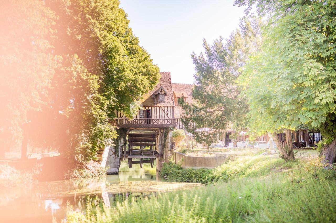 Vieille maison à colombages surplombant un cours d'eau, entourée d'arbres et baignée d'une lumière douce.