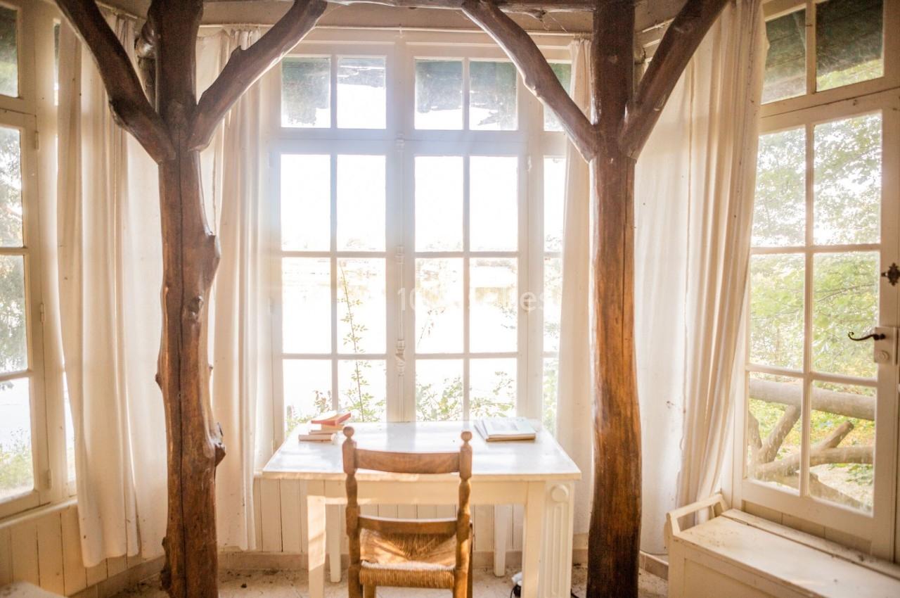 Bureau en bois clair avec une chaise, encadré par des troncs d'arbres, devant une grande baie vitrée lumineuse.