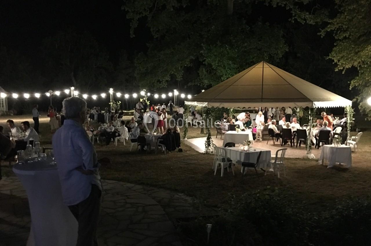 Vue nocturne d'une réception en plein air avec des invités assis sous un chapiteau éclairé par des guirlandes lumineuses.