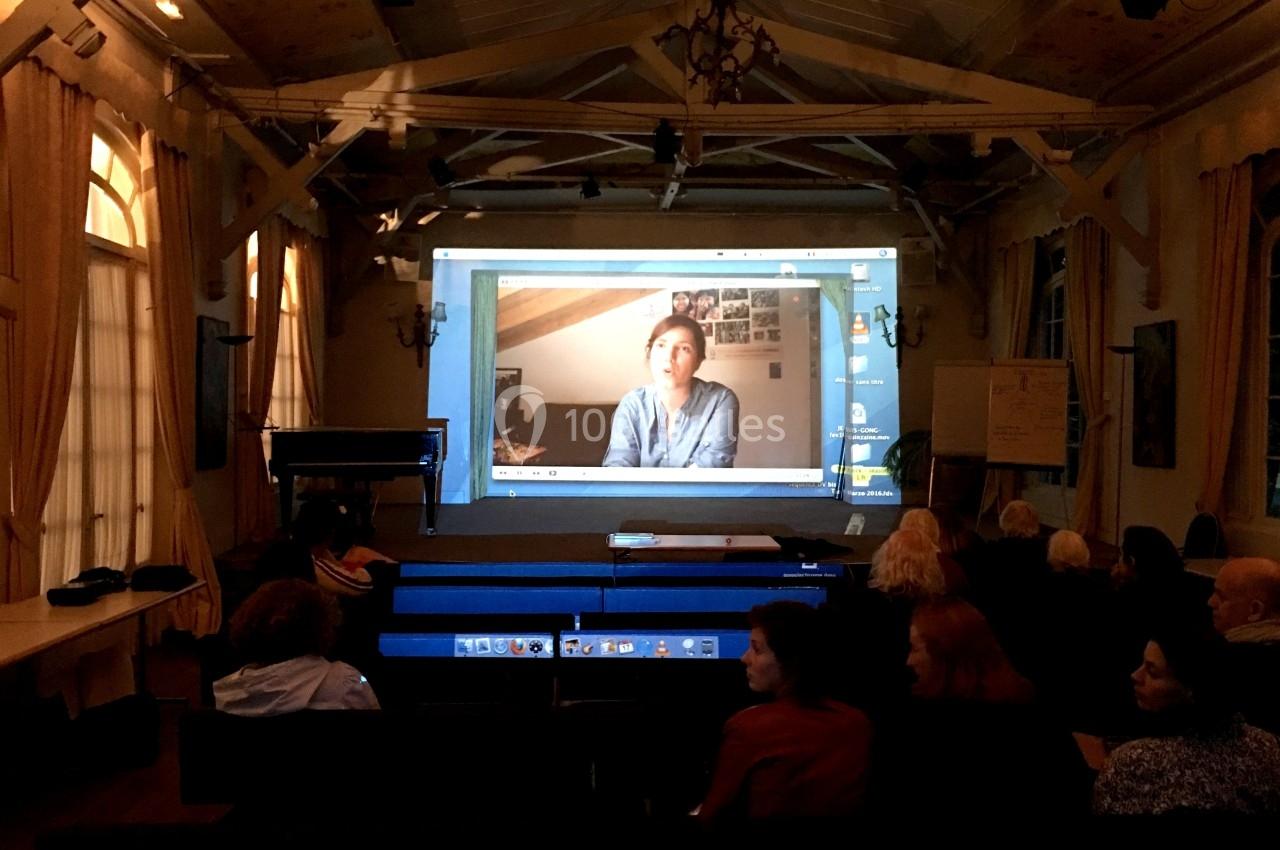 Projection d'une vidéo montrant une personne dans une salle sombre avec un public assis devant l'écran.