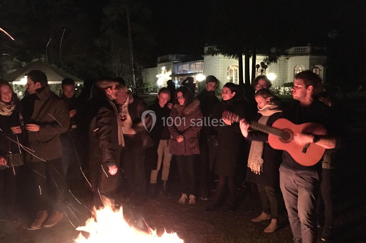 Un groupe de personnes debout autour d'un feu de camp, dont une jouant de la guitare, en soirée devant un bâtiment éclairé.
