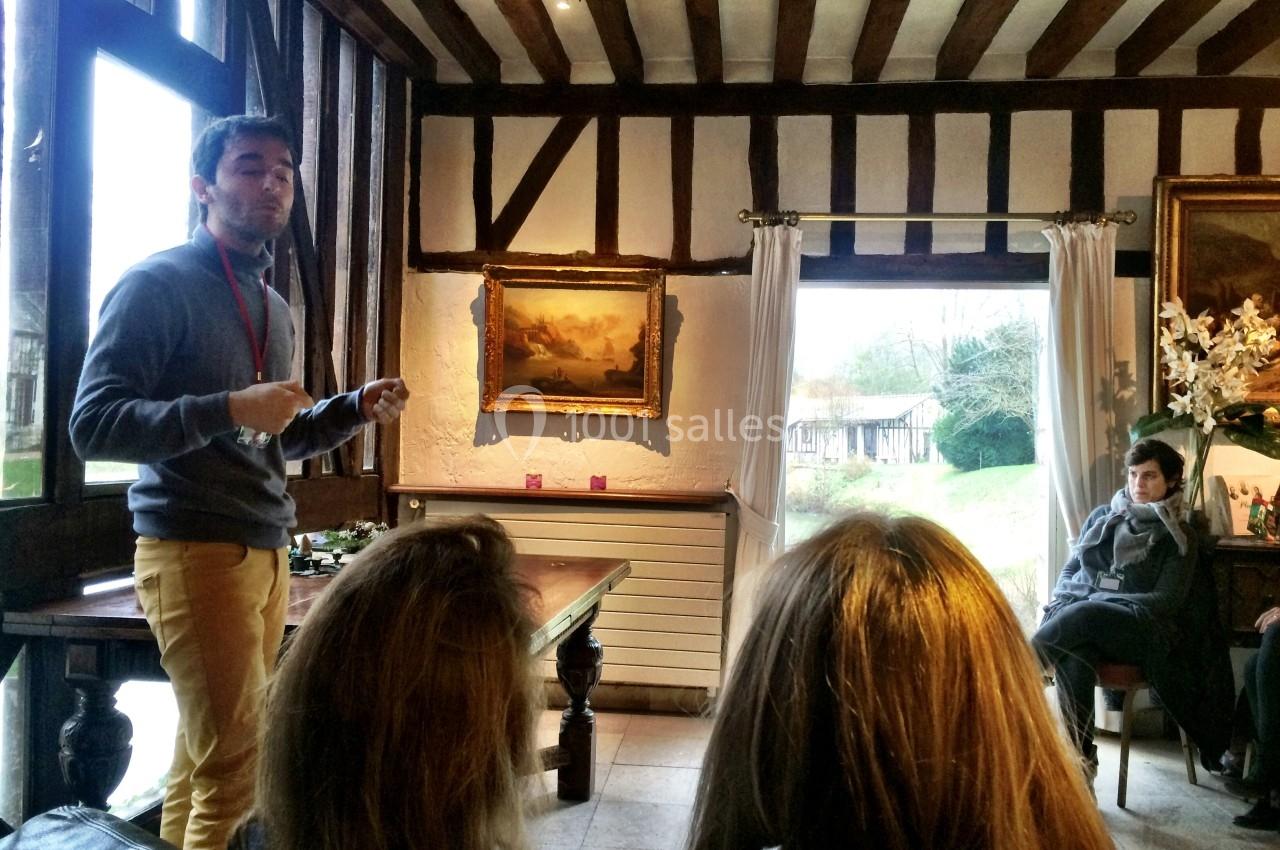 Un homme présente devant un petit groupe dans une salle avec poutres apparentes et une grande fenêtre donnant sur un jardin.