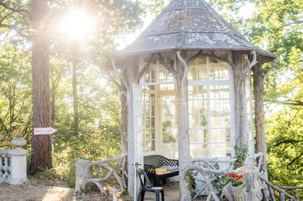 Petit pavillon en bois et verre entouré de végétation, avec une chaise noire devant et un panneau indiquant ’Moulin’.