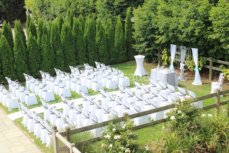 Chaises blanches alignées en extérieur pour une cérémonie, entourées de verdure et décorées de rubans argentés.
