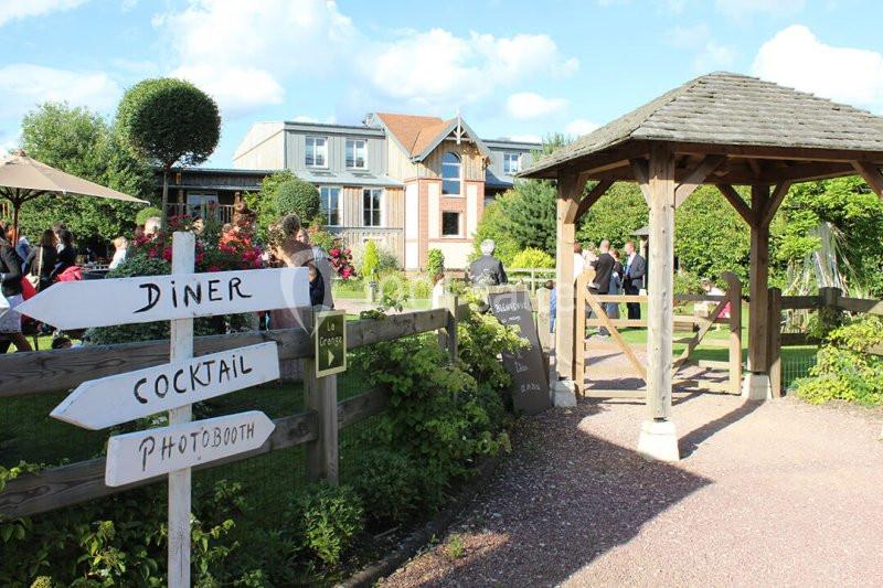 Entrée d'un jardin avec un panneau indiquant ’Dîner’, ’Cocktail’ et ’Photobooth’, près d'une pergola en bois.
