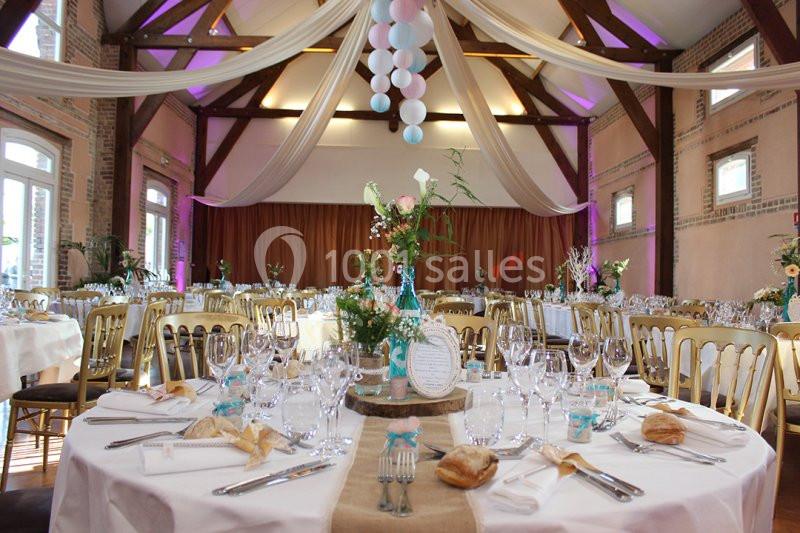 Salle de réception décorée pour un événement, avec tables dressées, chaises dorées et guirlandes suspendues.