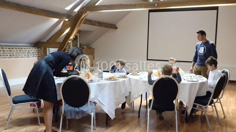 Des enfants assis autour de tables rondes dans une salle lumineuse, accompagnés de deux adultes.