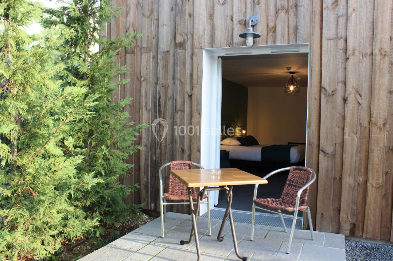 Terrasse avec table et chaises devant une porte ouverte donnant sur une chambre lumineuse, entourée de bois et de verdure.