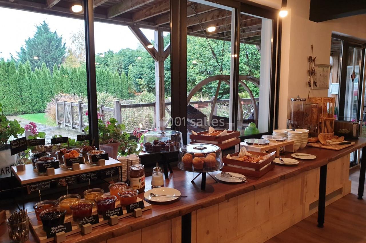 Buffet de petit-déjeuner avec viennoiseries, confitures, pains et boissons, près d'une baie vitrée donnant sur un jardin.