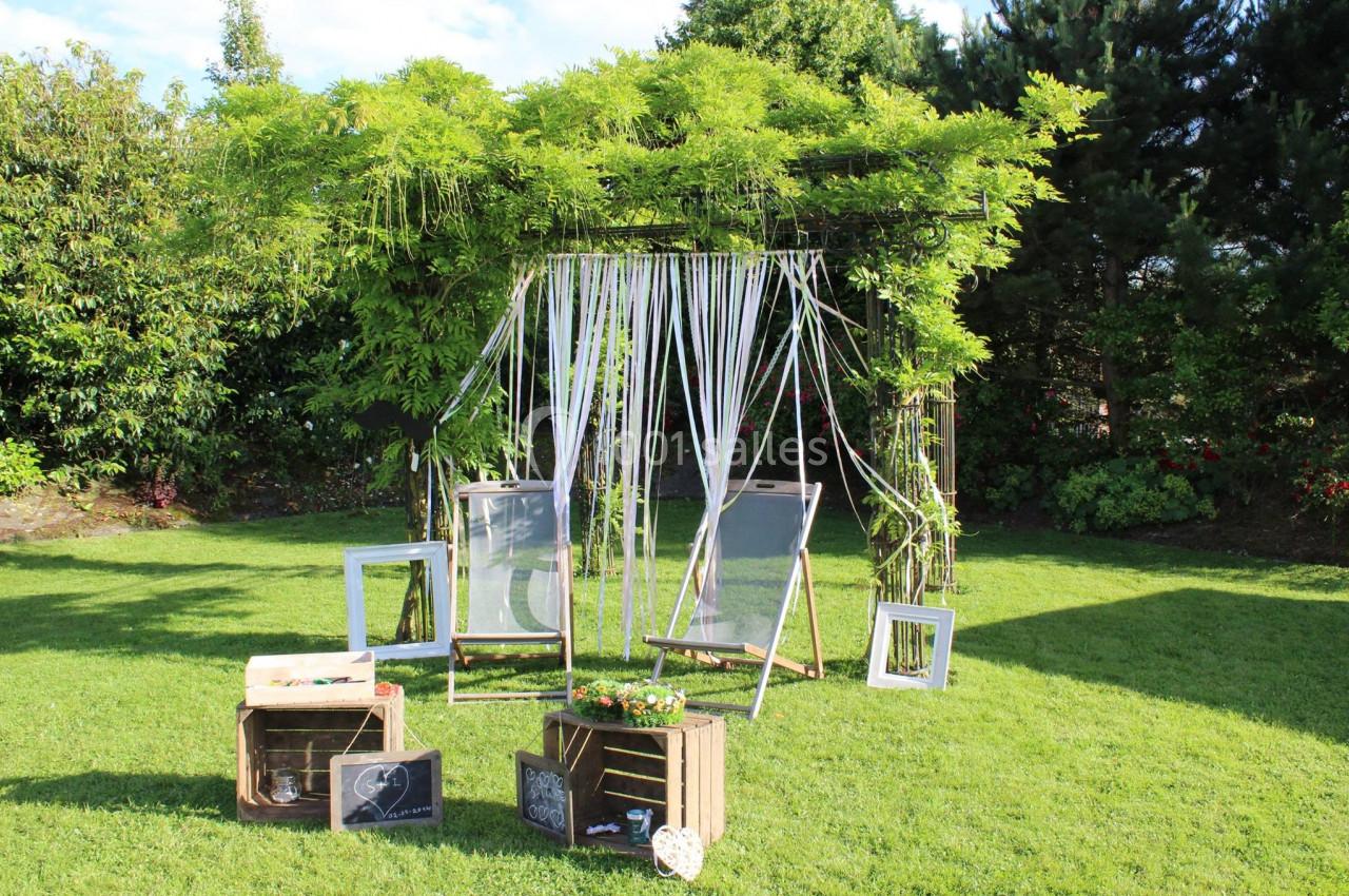 Deux chaises longues sous une pergola ornée de rubans et de verdure, entourées de caisses en bois et cadres décoratifs.
