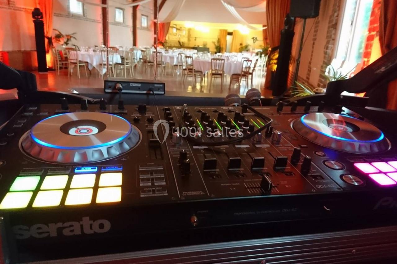 Table de mixage DJ avec platines lumineuses en premier plan, salle de réception décorée et tables dressées en arrière-plan.