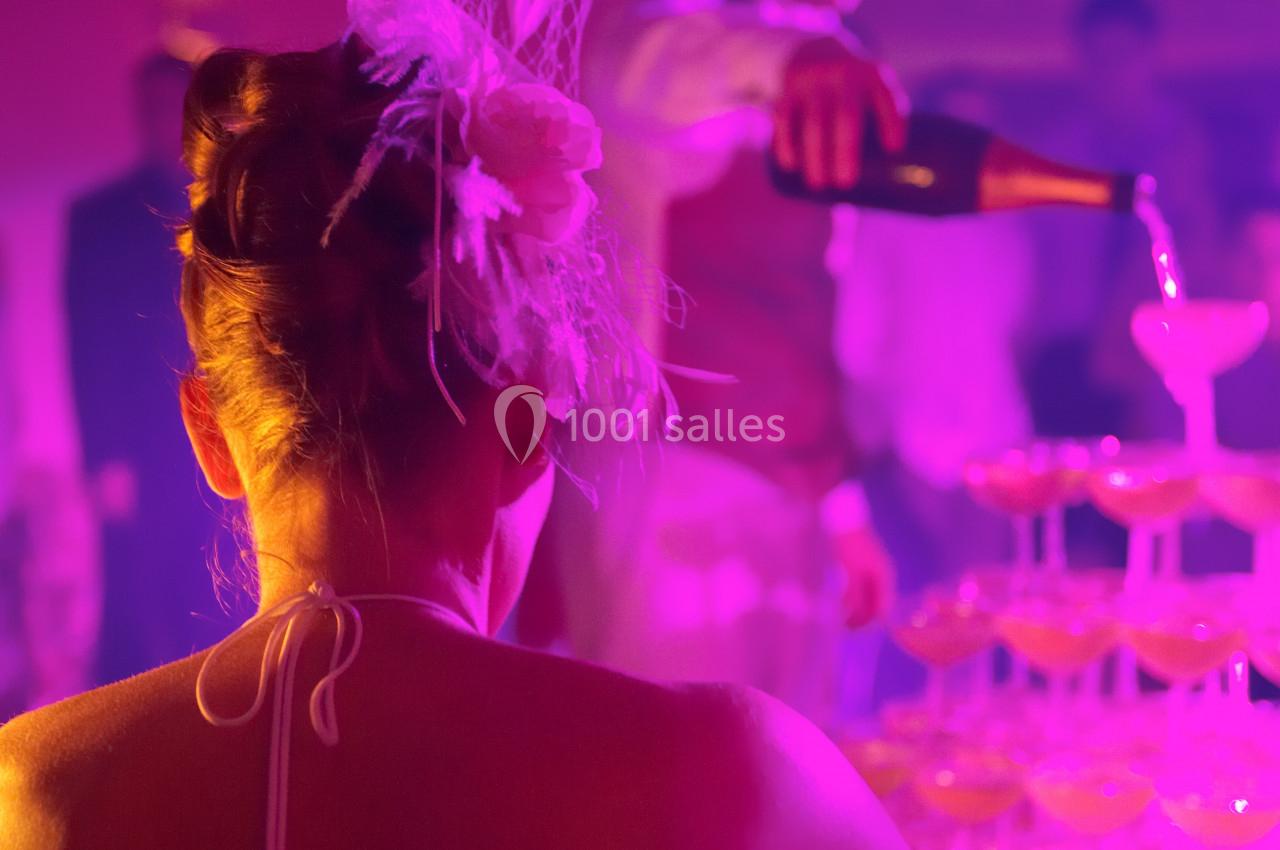 Une femme de dos observe un serveur verser du champagne dans une pyramide de coupes sous une lumière tamisée.