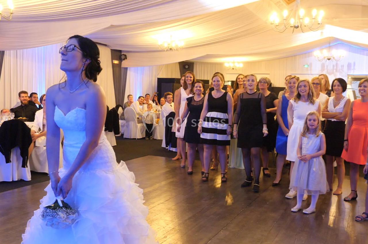 Une mariée lance son bouquet de fleurs à un groupe de femmes rassemblées dans une salle de réception décorée.