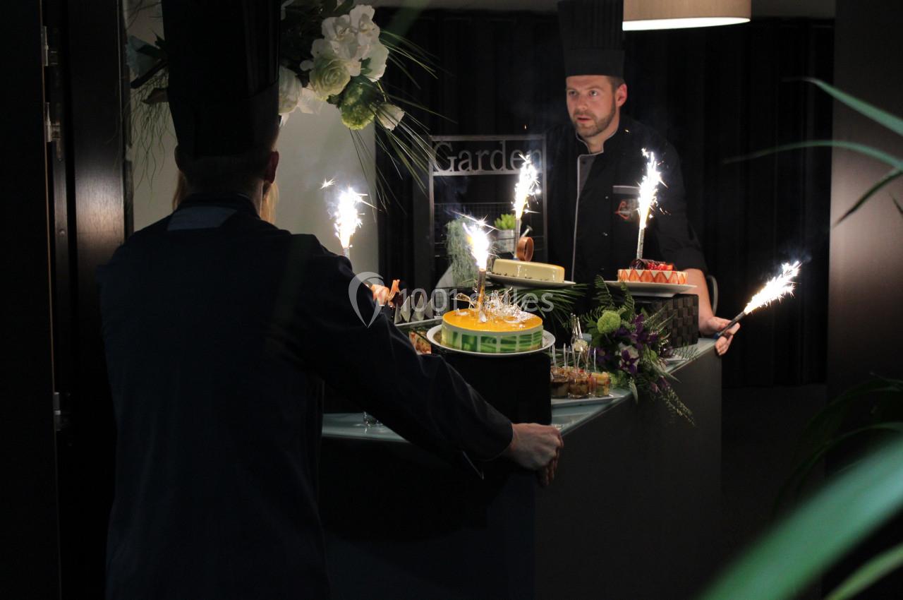 Deux chefs en tenue présentent des desserts avec des cierges magiques sur un comptoir décoré de fleurs.