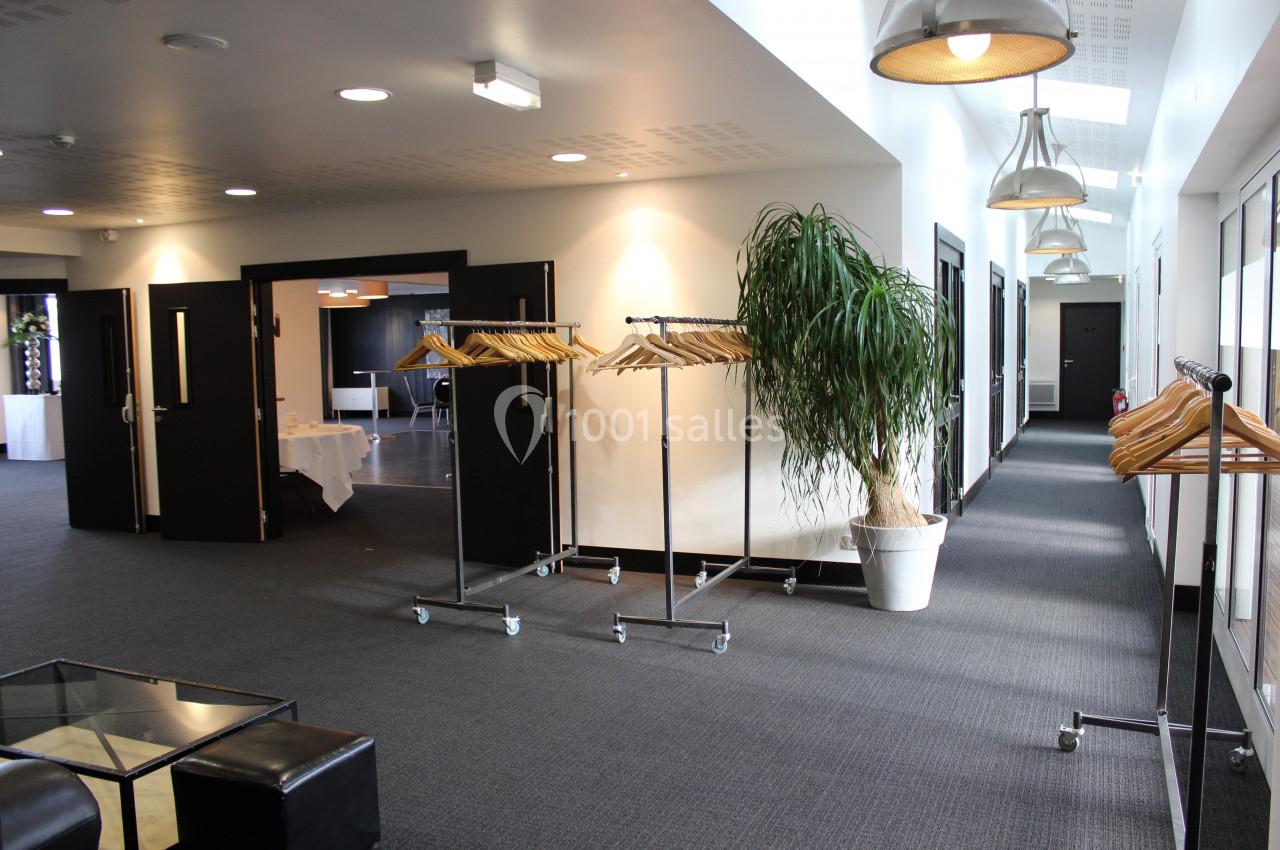 Portant à vêtements sur roulettes dans un couloir lumineux avec sol moquetté, plantes en pot et portes ouvertes.