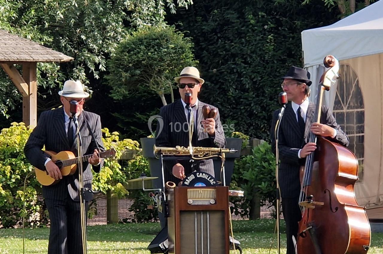 Trois musiciens en costume jouent de la guitare, du saxophone et de la contrebasse dans un jardin en plein air.
