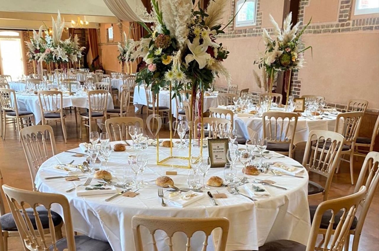 Salle de réception décorée avec des tables rondes dressées, ornées de centres de table floraux et de chaises en bois clair.