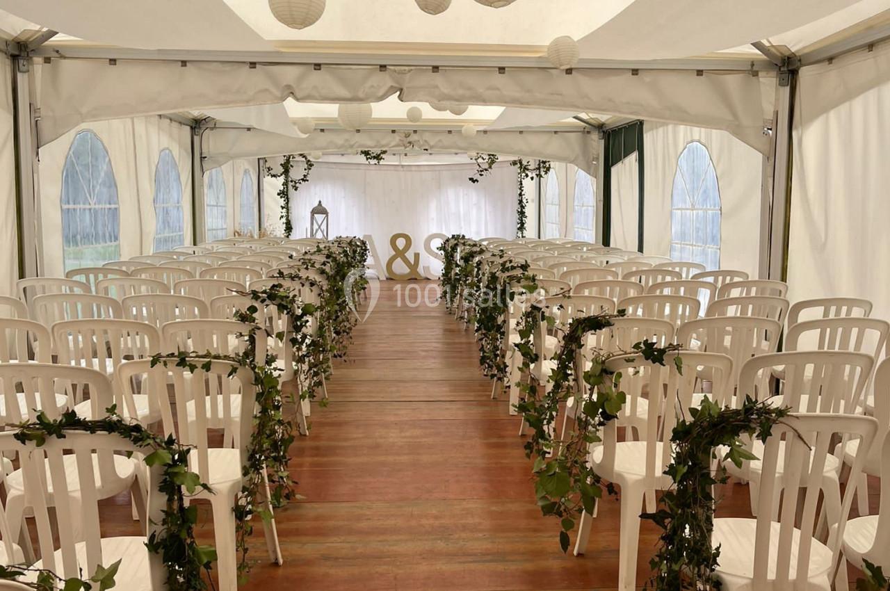 Allée centrale d'une tente de mariage décorée de guirlandes de lierre, avec des chaises blanches et des lanternes suspendues.