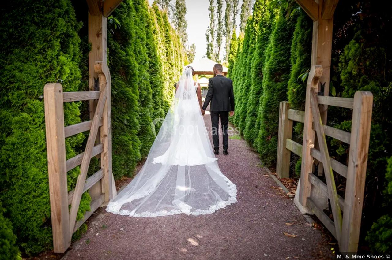 Un couple de mariés marche main dans la main sur un chemin bordé de haies verdoyantes.