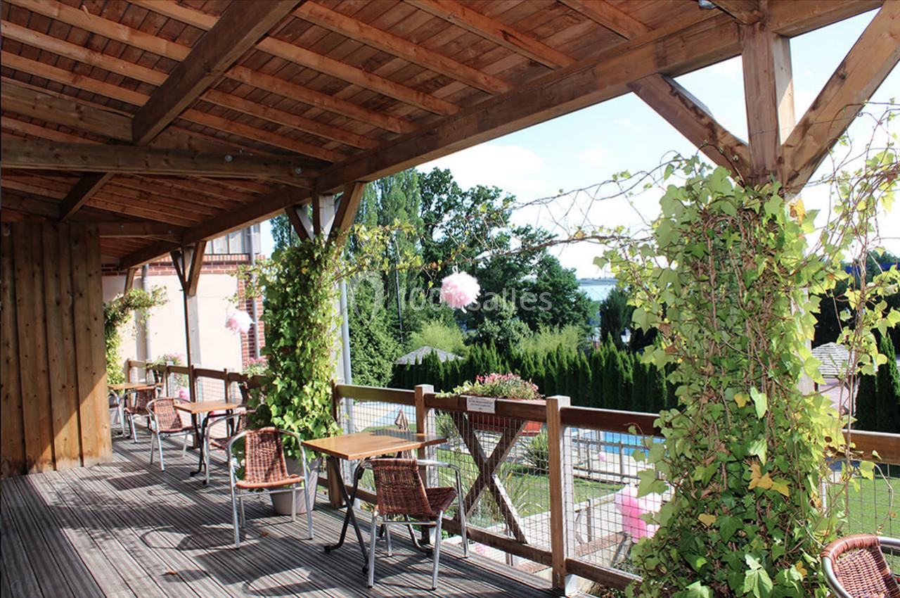 Terrasse en bois couverte avec tables et chaises, entourée de plantes grimpantes, offrant une vue sur un jardin verdoyant.