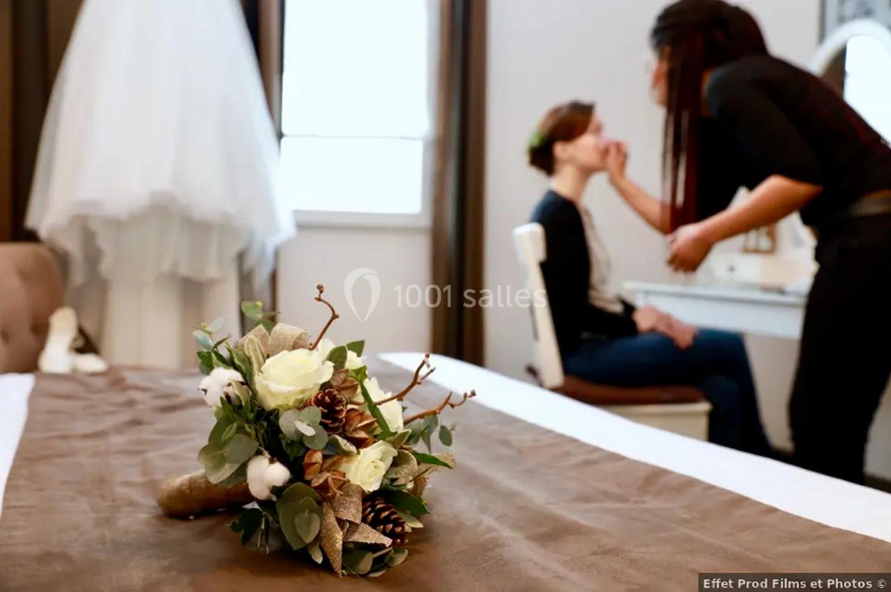 Un bouquet de fleurs posé sur un lit au premier plan, une maquilleuse prépare une femme en arrière-plan.