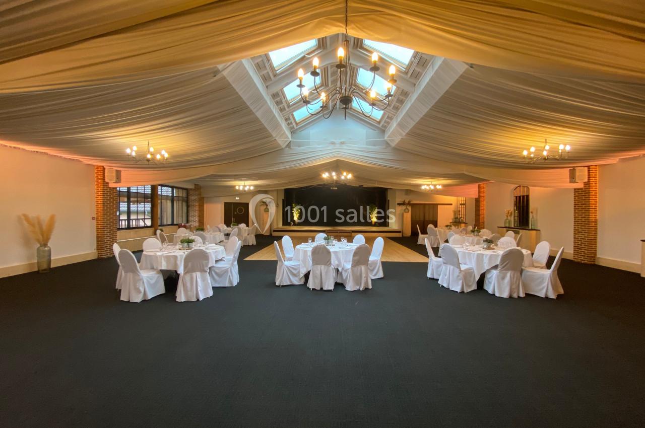Salle de réception spacieuse avec tables rondes dressées de nappes blanches, éclairée par des lustres et une verrière.