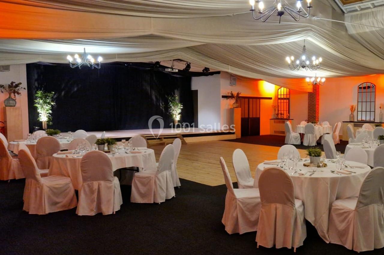Salle de réception décorée avec des tables rondes dressées, des chaises blanches et une scène en arrière-plan.