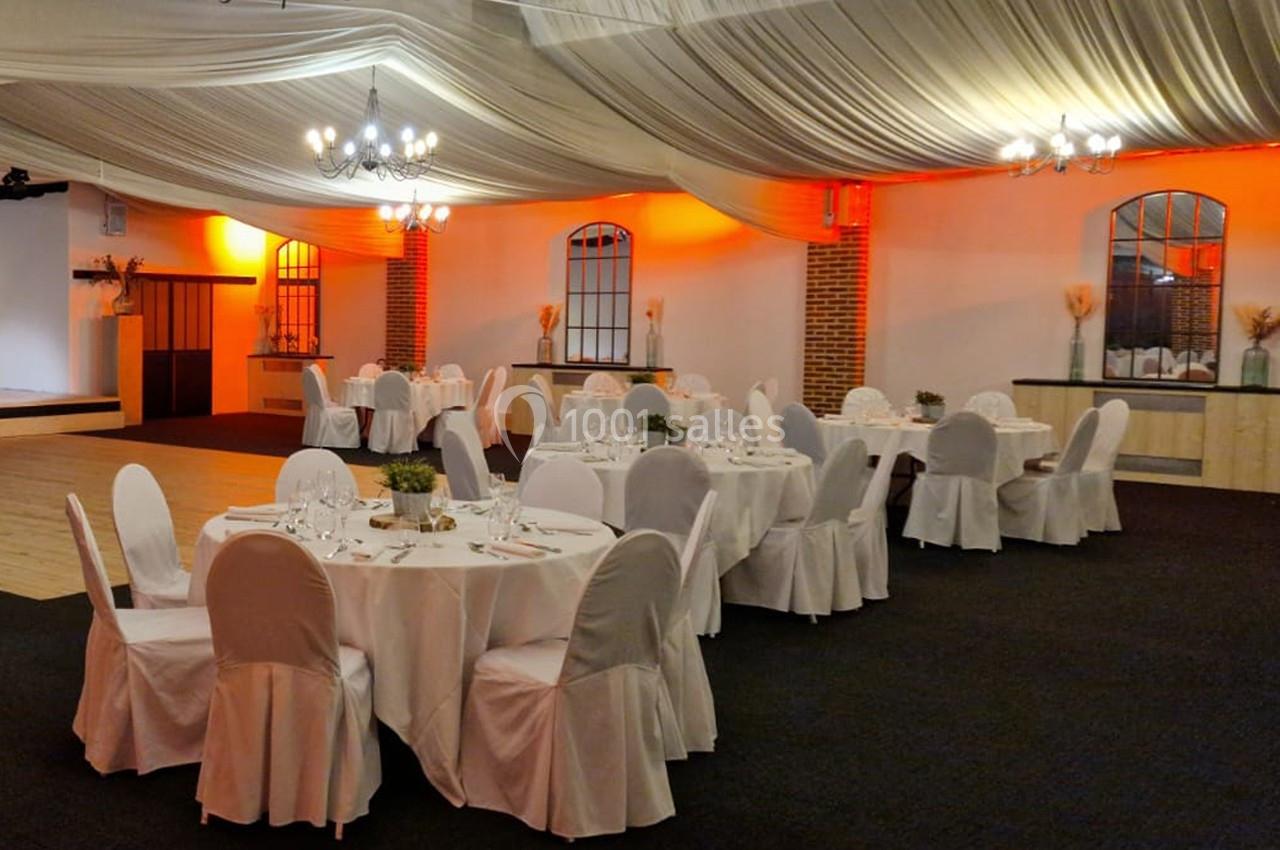 Salle de réception décorée avec des tables rondes dressées, chaises blanches et éclairage chaleureux sous un plafond drapé.