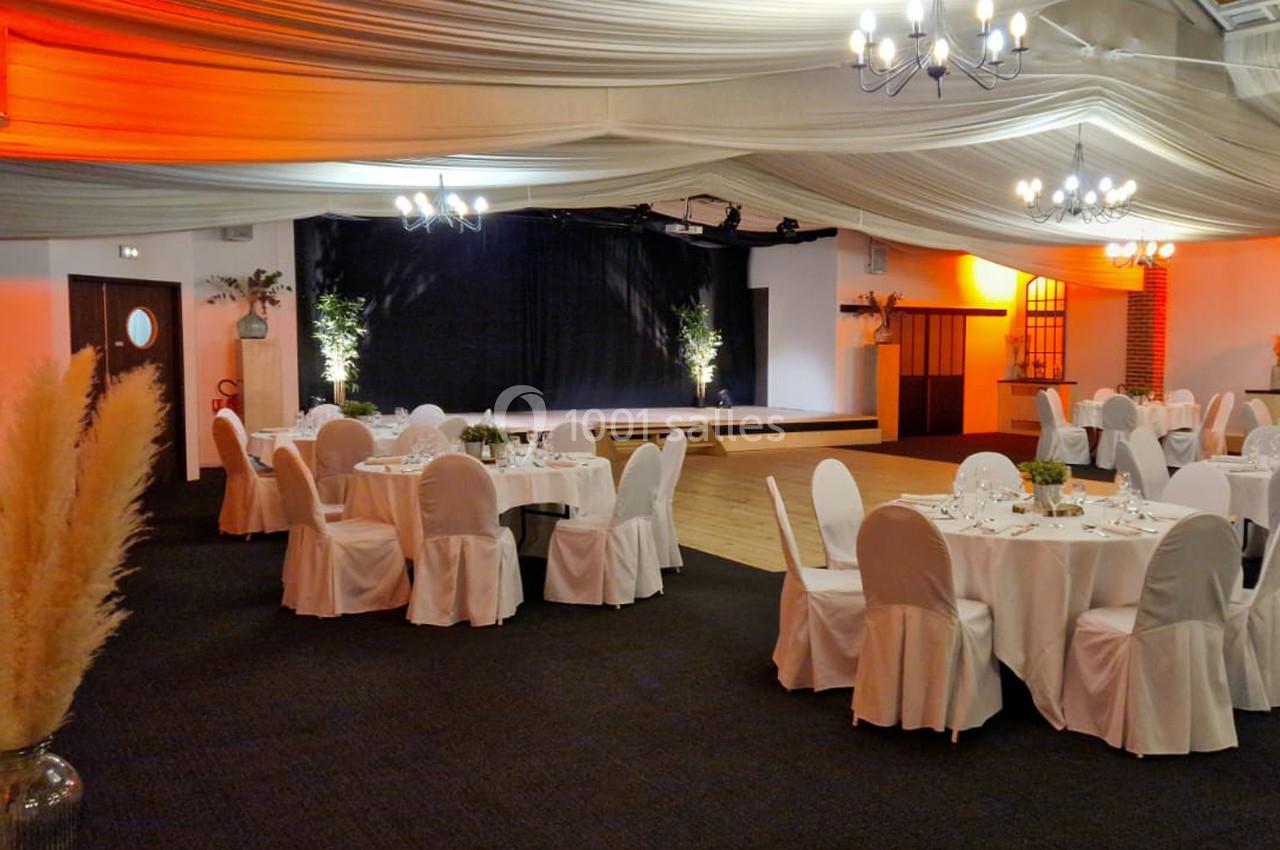 Salle de réception décorée avec des tables rondes dressées, des chaises blanches et une scène en arrière-plan.