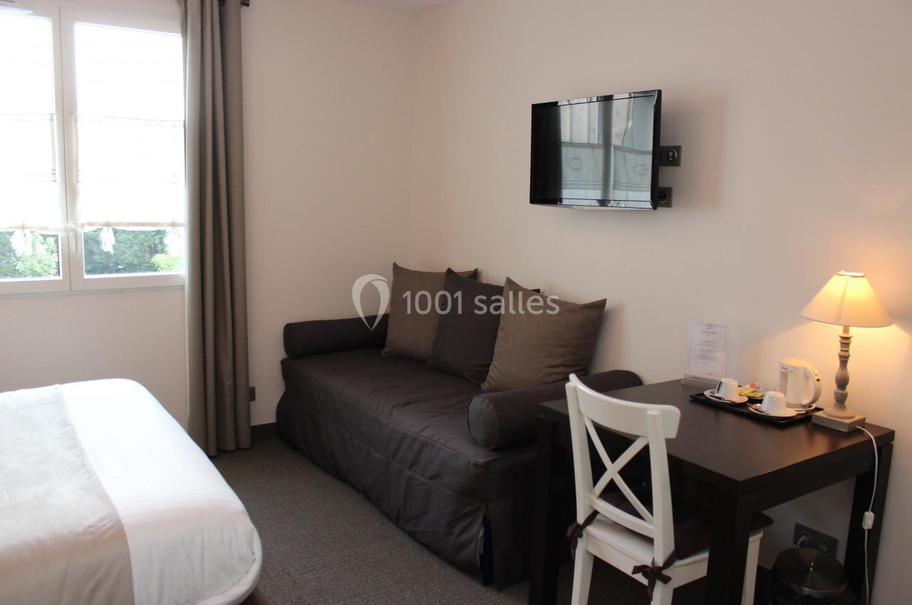 Chambre lumineuse avec lit double, canapé, bureau avec chaise, télévision murale et fenêtre donnant sur l'extérieur.