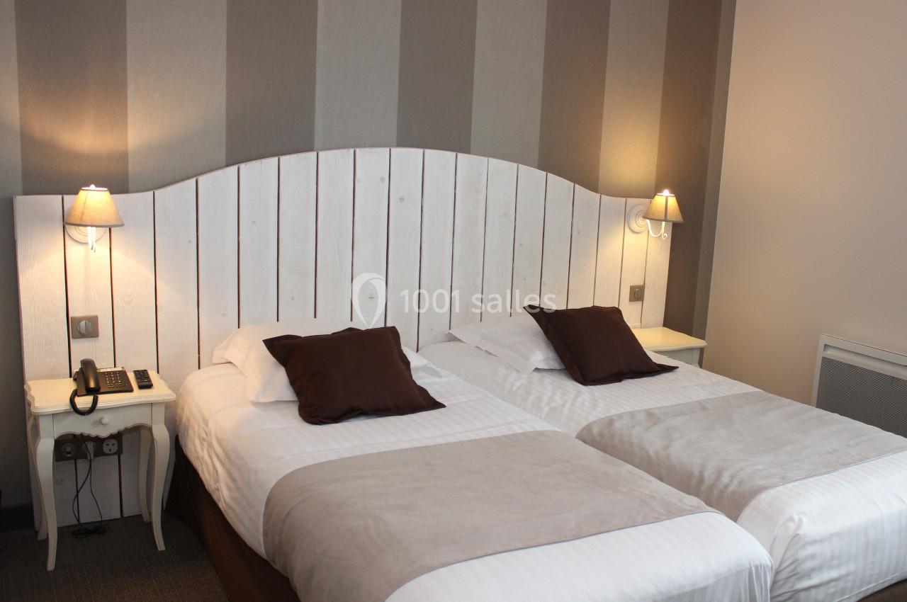 Deux lits simples avec linge blanc et coussins marron, éclairés par deux lampes murales dans une chambre sobre.