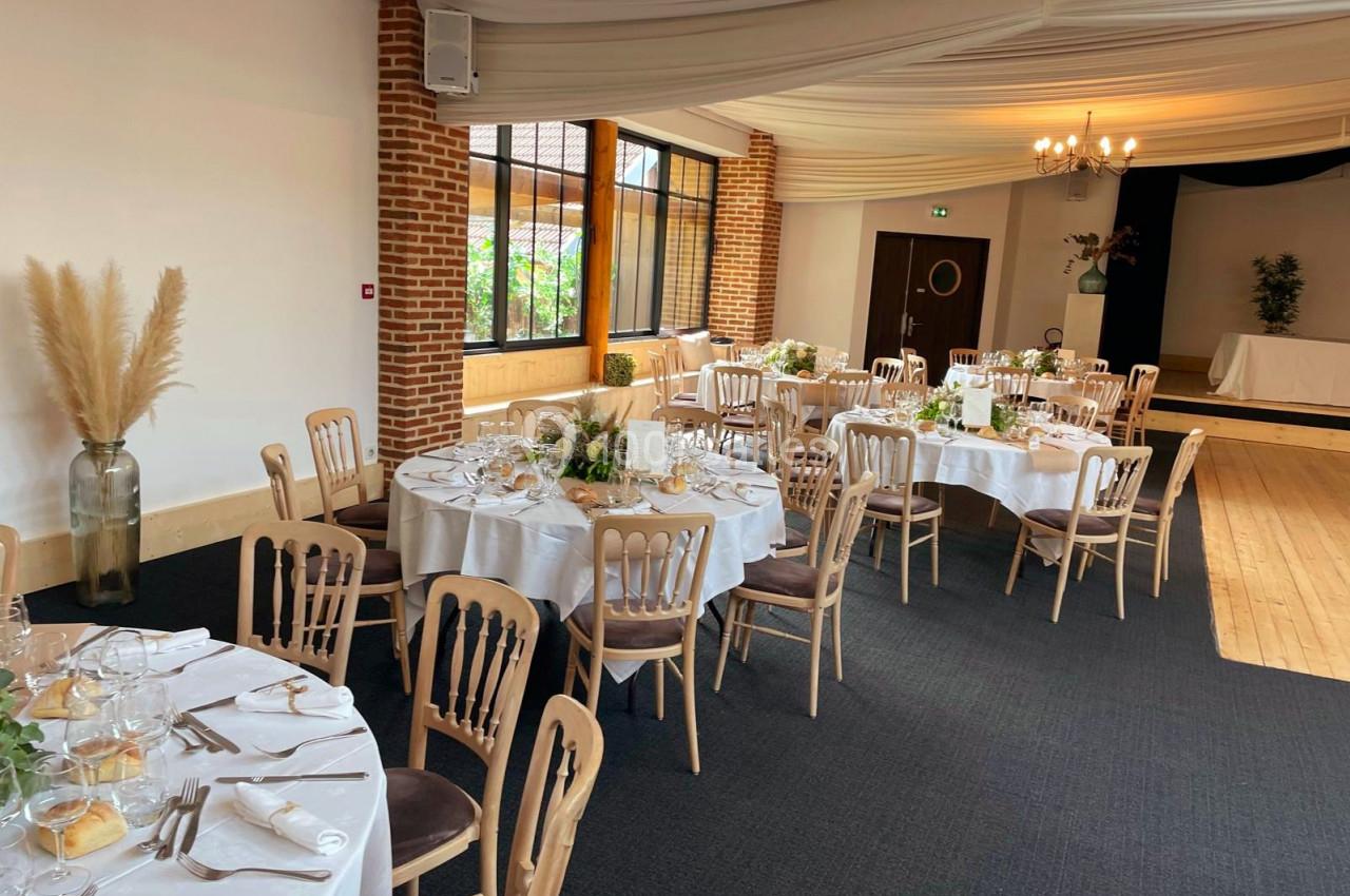 Salle de réception décorée avec des tables rondes dressées, nappes blanches, chaises en bois et végétation en centre de…