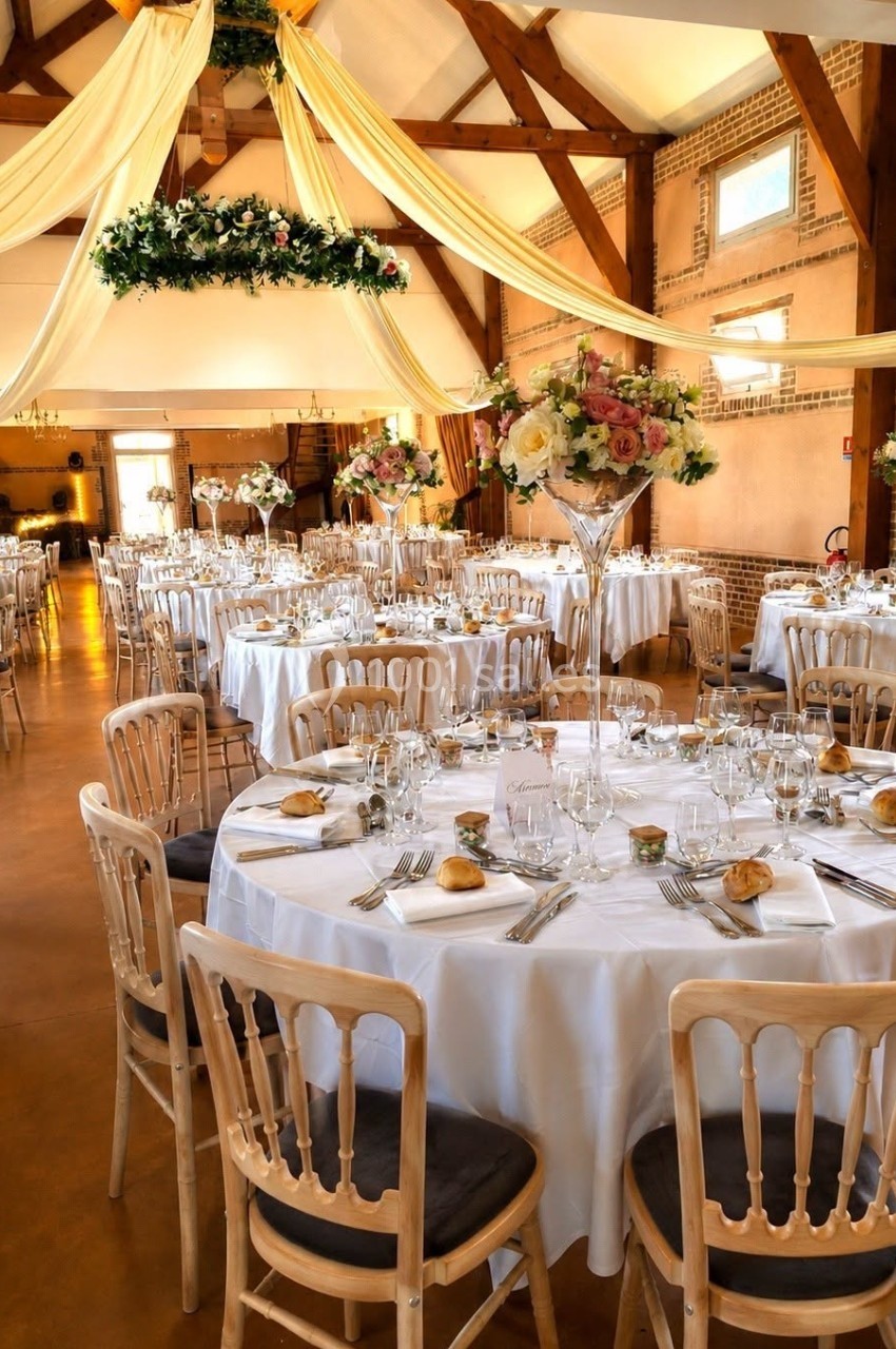 Salle de réception décorée avec des tables rondes dressées, nappes blanches, fleurs et chaises en bois clair.